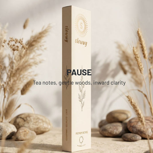 A tall rectangular box of slowwy brand incense stands upright on a stone surface with dried grasses and smooth stones around it. The box is warm off-white/cream with gold text and botanical line art.