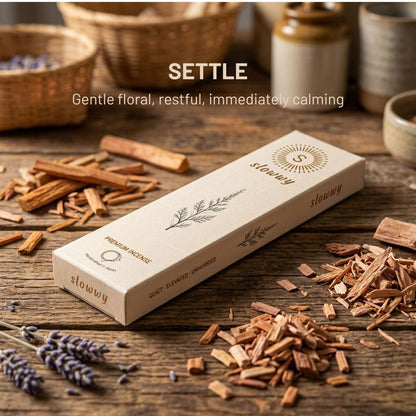 A rectangular box of slowwy incense boxes sits on a rustic wooden table surrounded by scattered wood shavings, cinnamon sticks, lavender sprigs, and blurred jars in the background. White text above reads 'SETTLE' Gentle floral, restful, immediately calming.