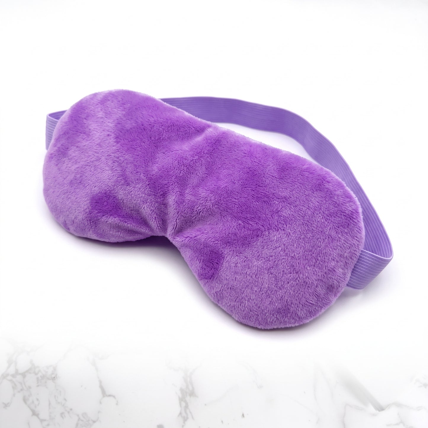 A plush purple sleep mask with an adjustable elastic strap, displayed on a white marble-patterned surface.