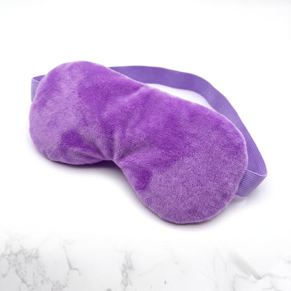 A plush purple sleep mask with an adjustable elastic strap, displayed on a white marble-patterned surface.