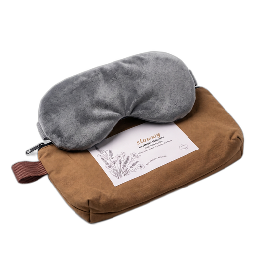 A gray plush sleep mask resting on top of a brown travel pillow set with a printed insert card, photographed on a clean white background.