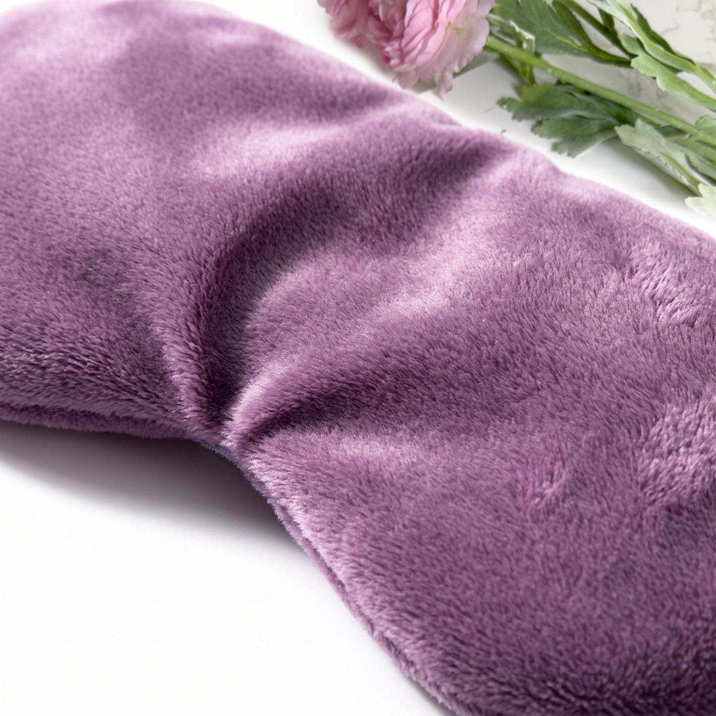 A close-up of a plush, soft purple eye mask with a velvet-like texture, styled with pink flowers and green leaves in the top right corner on a white background.