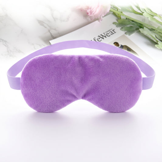A plush purple sleep mask with an elastic strap, displayed on a clean white surface with a magazine and flowers in the background.