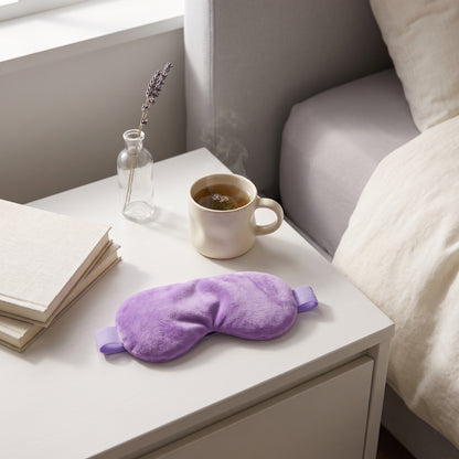 A purple plush eye mask rests on a white bedside table next to a mug of tea, a small glass vase with lavender, and a stack of books, with a bed visible in the background.