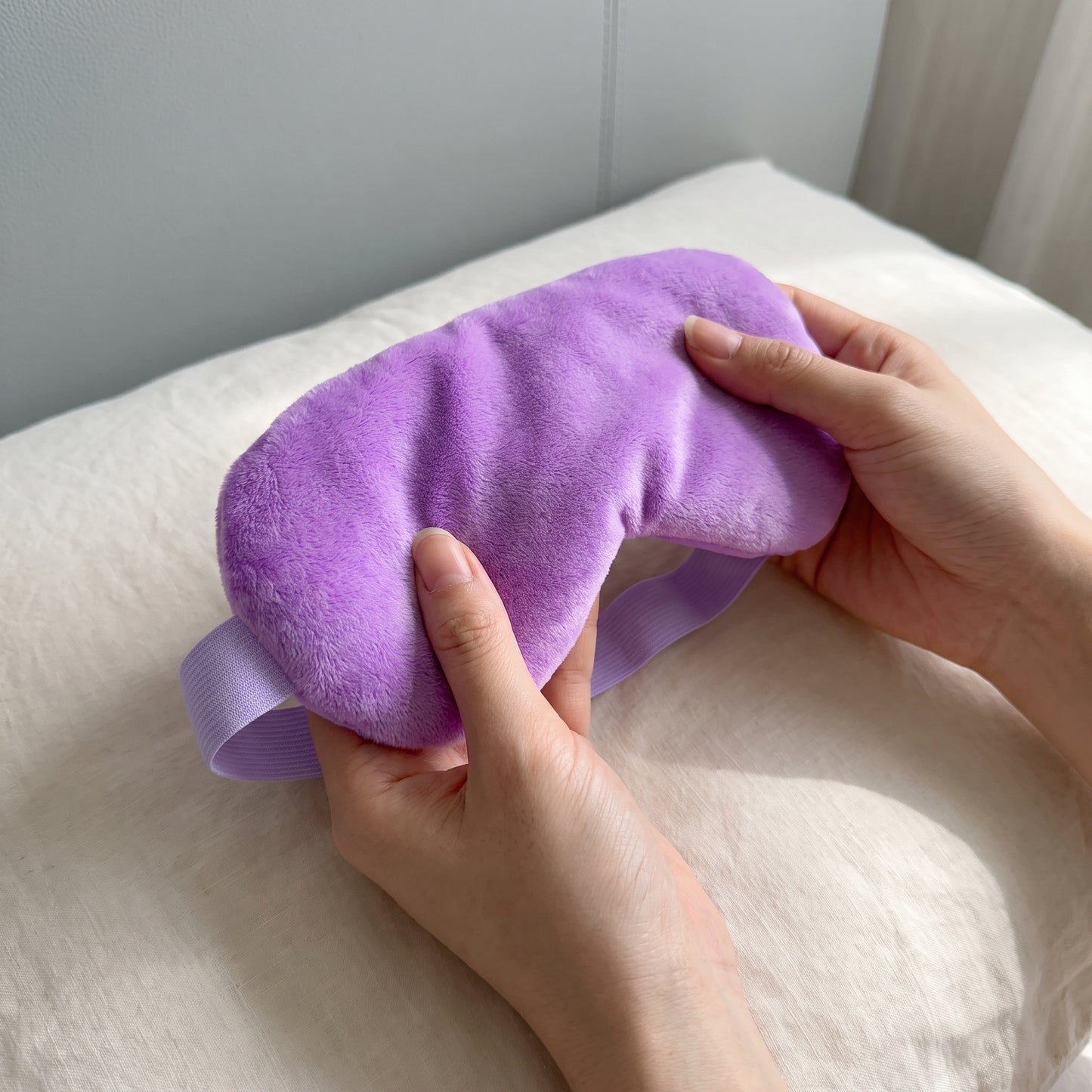 A purple plush eye mask being held by two hands over a white pillow, with a matching elastic strap visible underneath.