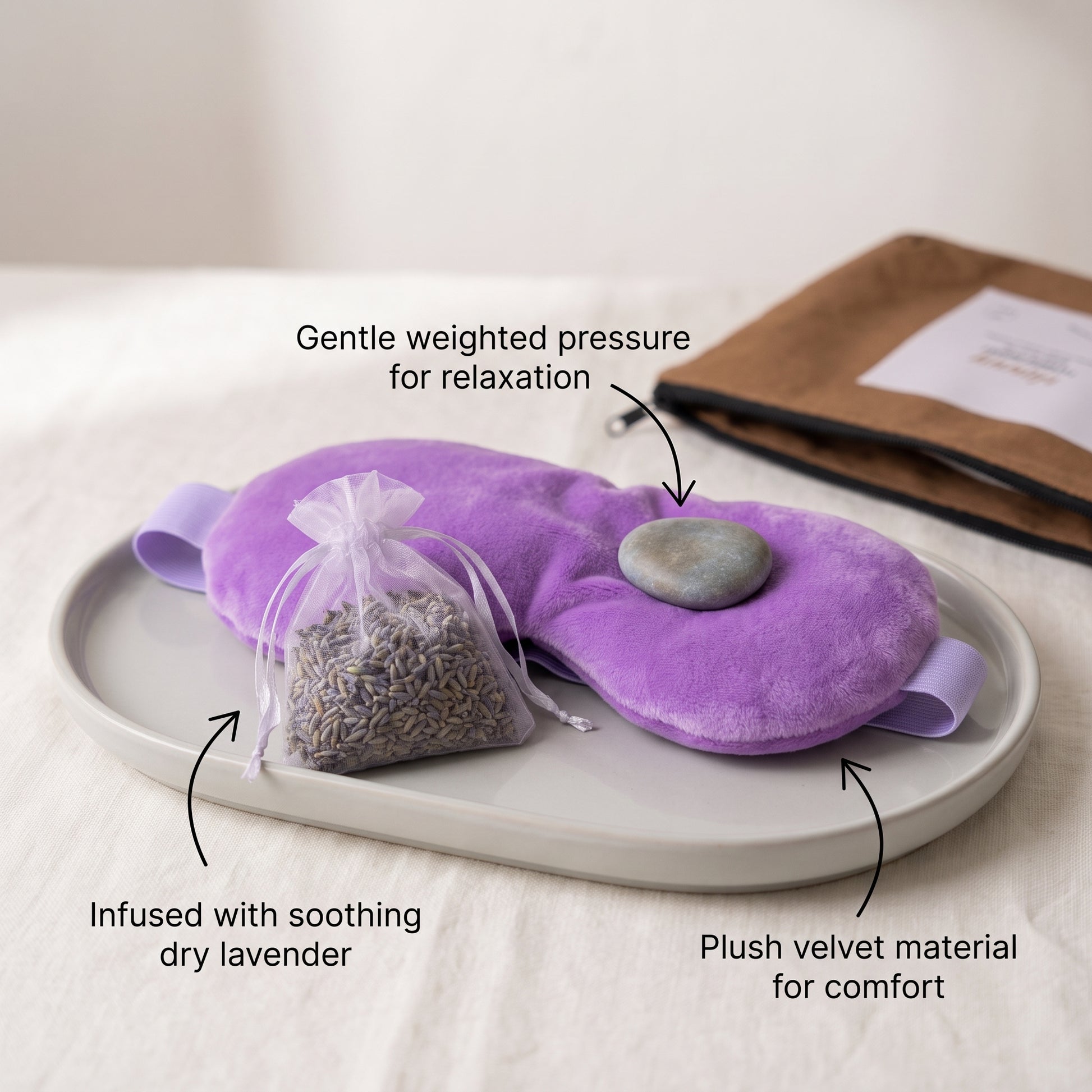 A purple plush eye mask set on a light gray tray, with a small sachet of dried lavender and a smooth stone. Text labels describe the features: gentle weighted pressure for relaxation, infused with soothing dry lavender, and plush velvet material for comfort.
