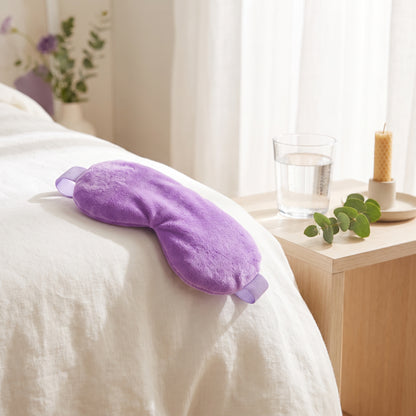 A purple plush eye mask resting on a white bed in a softly lit bedroom, with a bedside table, glass of water, candle, and greenery nearby.