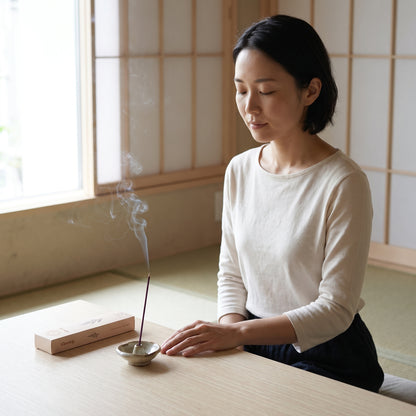 A woman in a white long-sleeve top sits at a low wooden table in a Japanese-style room, holding a incense stick over a small bowl. A rectangular slowwy incense box rests beside the bowl, with smoke rising.