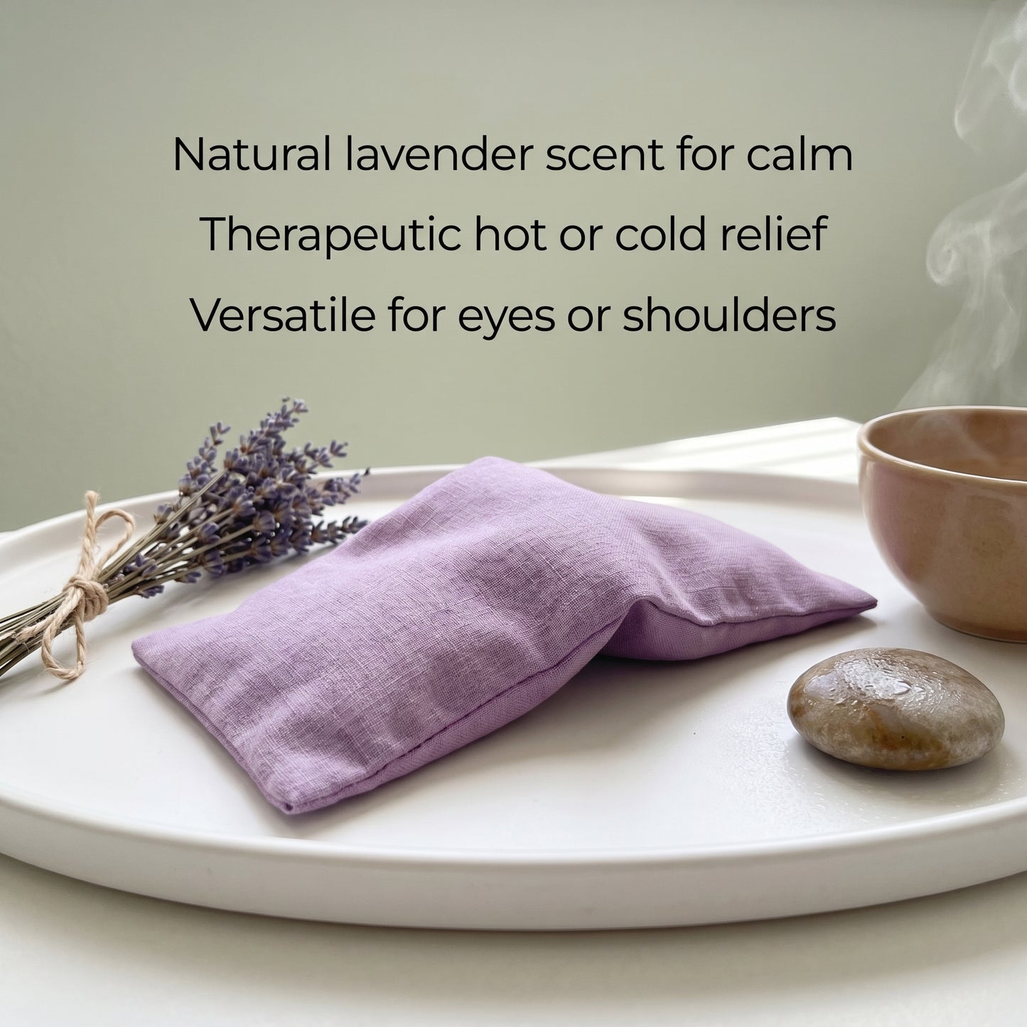 A lavender-scented fabric eye pillow in a soft purple shade, resting on a white tray alongside dried lavender, a small stone, and a ceramic cup. Text on the image reads: 'Natural lavender scent for calm, Therapeutic hot or cold relief, Versatile for eyes or shoulders'.