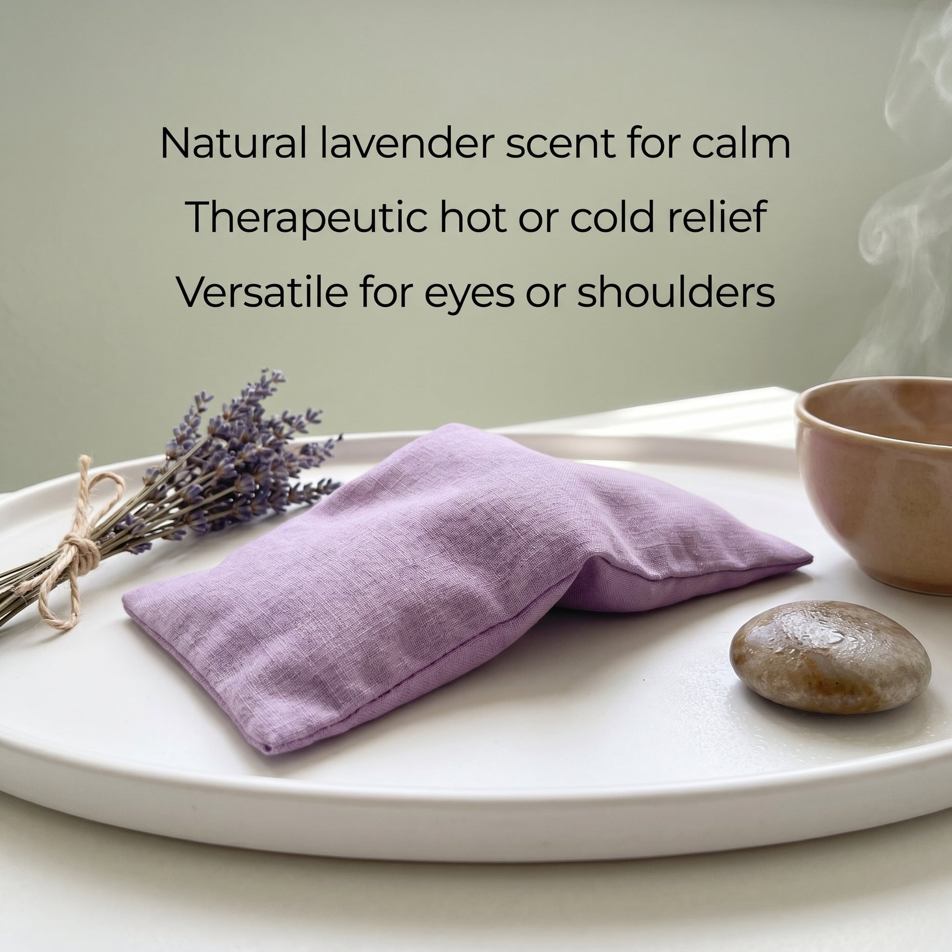 A lavender-scented fabric eye pillow in a soft purple shade, resting on a white tray alongside dried lavender, a small stone, and a ceramic cup. Text on the image reads: 'Natural lavender scent for calm, Therapeutic hot or cold relief, Versatile for eyes or shoulders'.